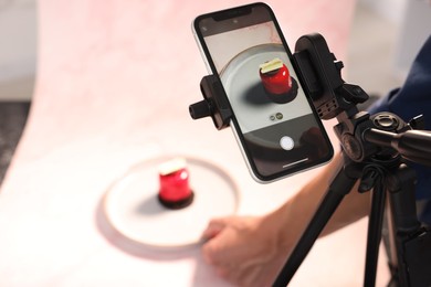 Photo of Man creating composition with tasty dessert for photoshoot at table in studio, focus on smartphone and tripod. Professional food photography