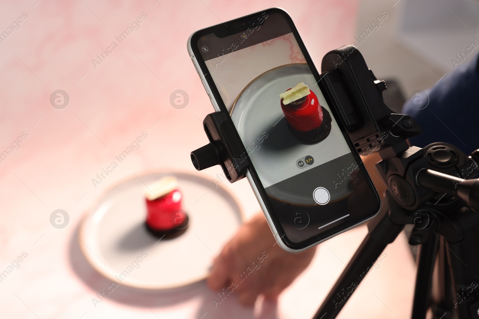 Photo of Man creating composition with tasty dessert for photoshoot at table in studio, focus on smartphone and tripod. Professional food photography