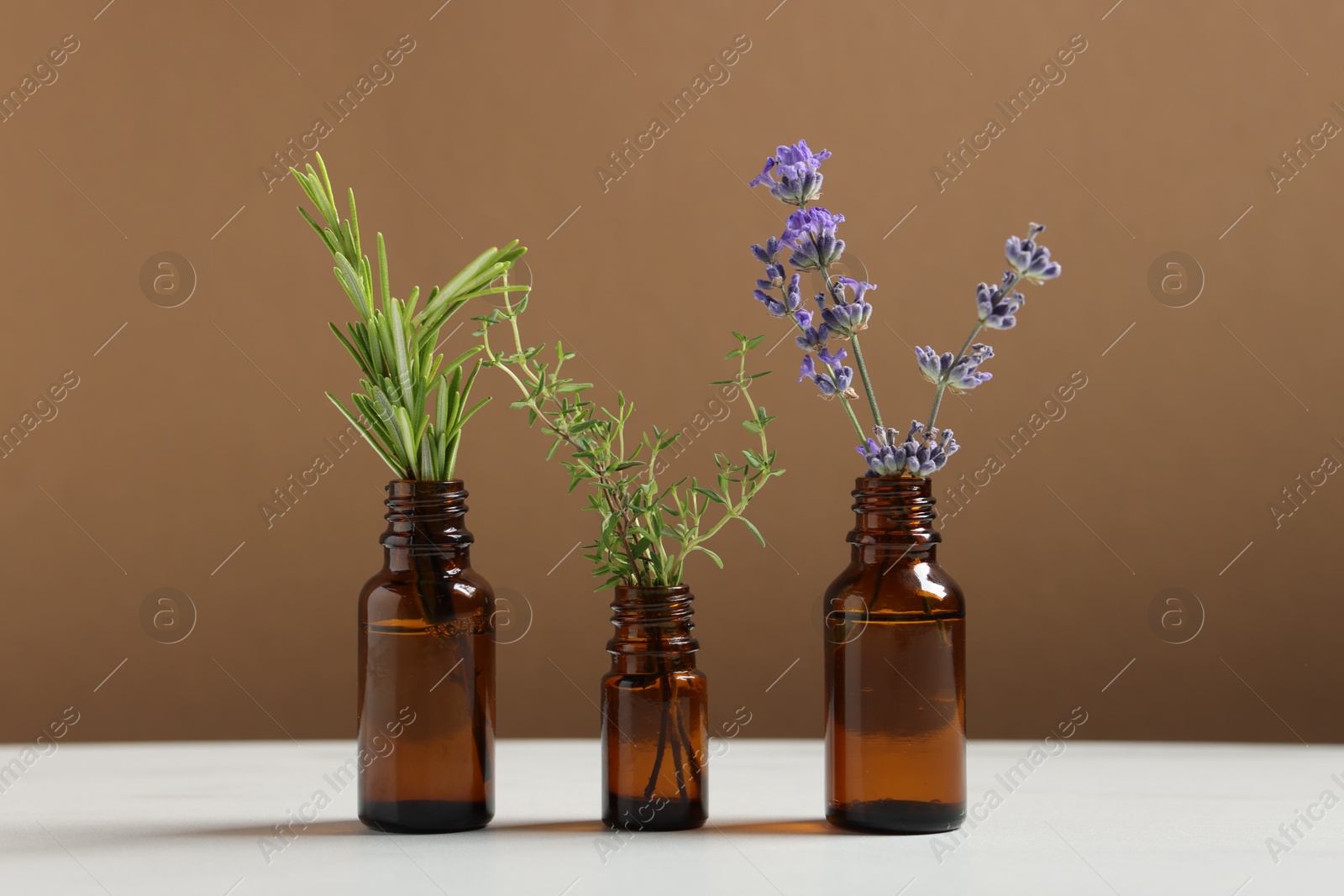 Photo of Different herbs in glass bottles of essential oils on white table against brown background