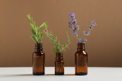 Photo of Different herbs in glass bottles of essential oils on white table against brown background