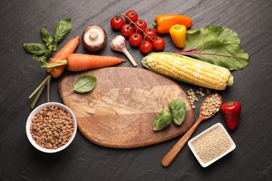 Photo of Healthy vegetarian food. Empty board surrounded by different products on dark textured table, flat lay