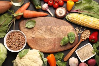 Photo of Healthy vegetarian food. Empty board surrounded by different products on dark textured table, flat lay