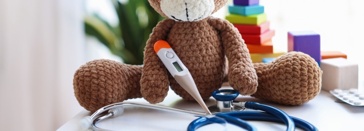 Photo of Pediatrician concept. Toy bear, thermometer and stethoscope on white table in clinic, closeup