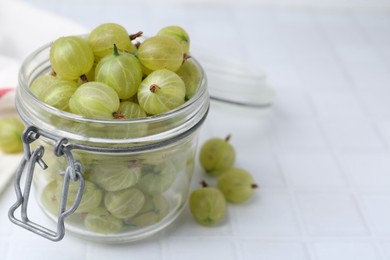 Photo of Fresh green gooseberries in jar on white tiled table, closeup. Space for text