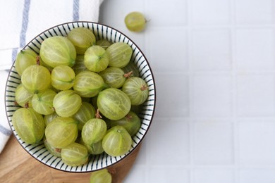 Photo of Fresh green gooseberries in bowl on white tiled table, top view. Space for text
