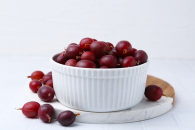 Photo of Ripe red gooseberries in bowl on white tiled table, closeup