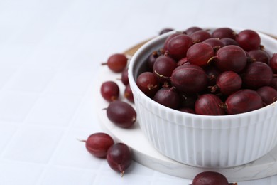 Photo of Ripe red gooseberries in bowl on white tiled table, closeup. Space for text