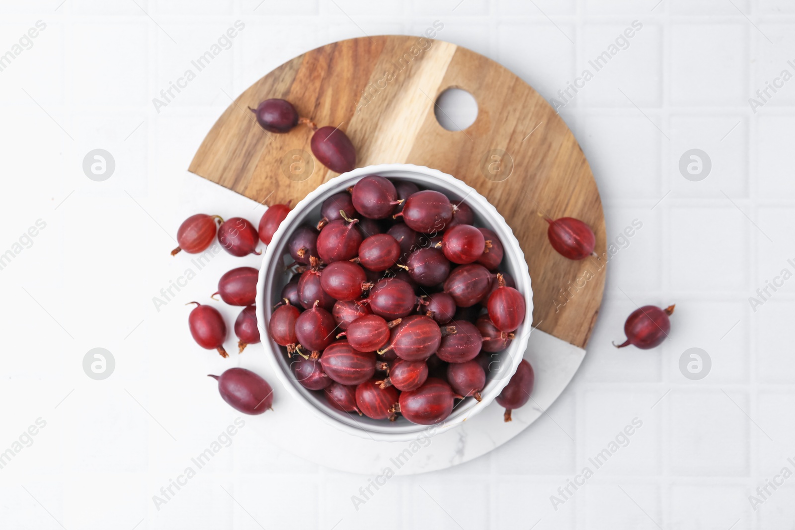 Ripe red gooseberries in bowl on white tiled table, flat lay Photo of Ripe red gooseberries in bowl on white tiled table, flat lay