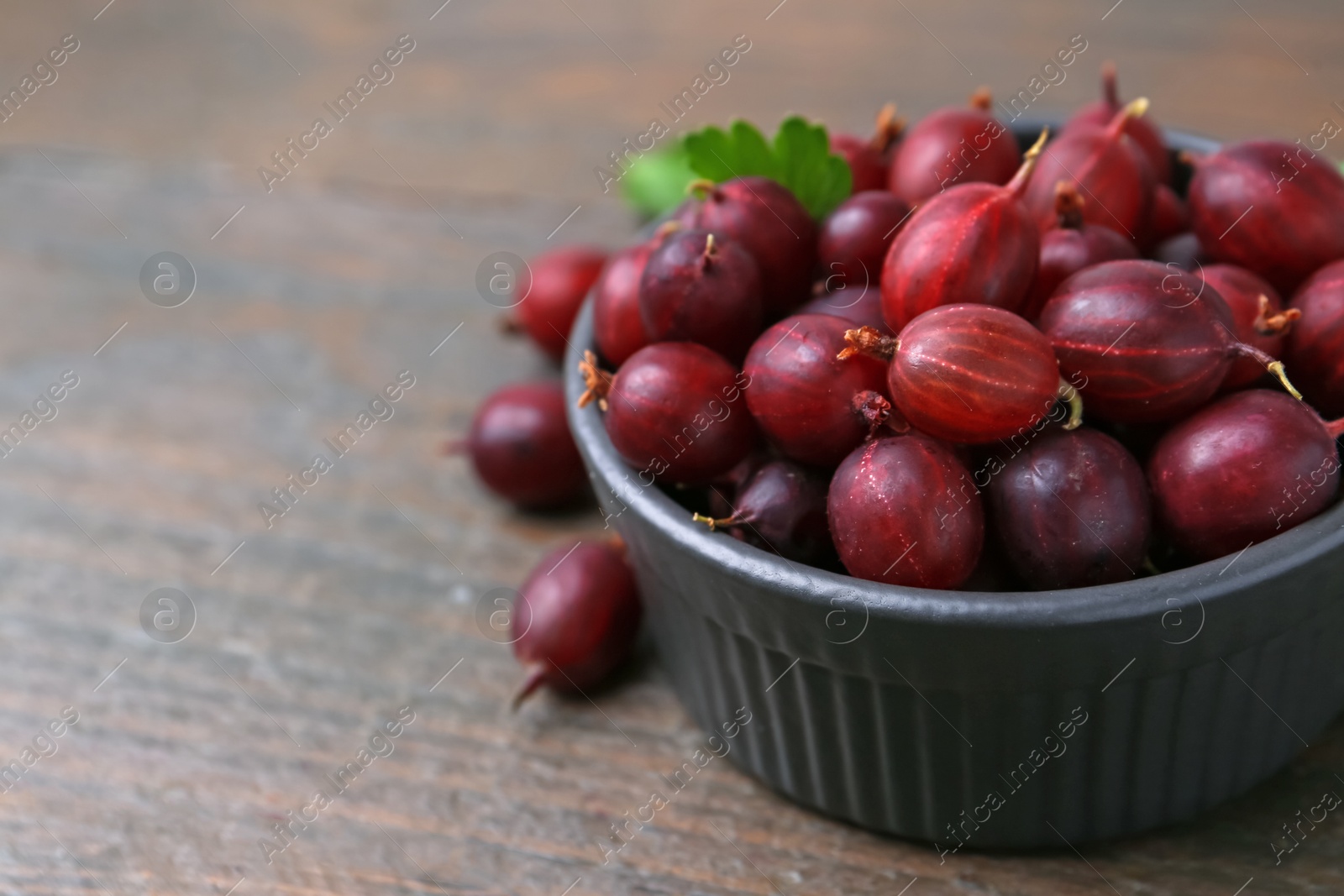 Photo of Ripe red gooseberries in bowl on wooden table, closeup. Space for text