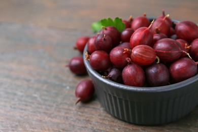 Photo of Ripe red gooseberries in bowl on wooden table, closeup. Space for text
