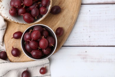 Photo of Ripe red gooseberries in bowl on white wooden table, flat lay. Space for text