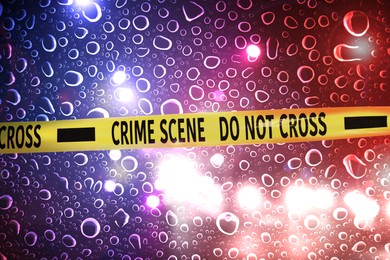 Image of Police yellow Crime Scene Do Not Cross tape. Red and blue lights, blurred view through wet glass