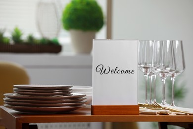 Image of Holder with word Welcome, clean dishware and cutlery on wooden table in restaurant