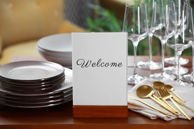 Image of Holder with word Welcome, clean dishware and cutlery on wooden table in restaurant