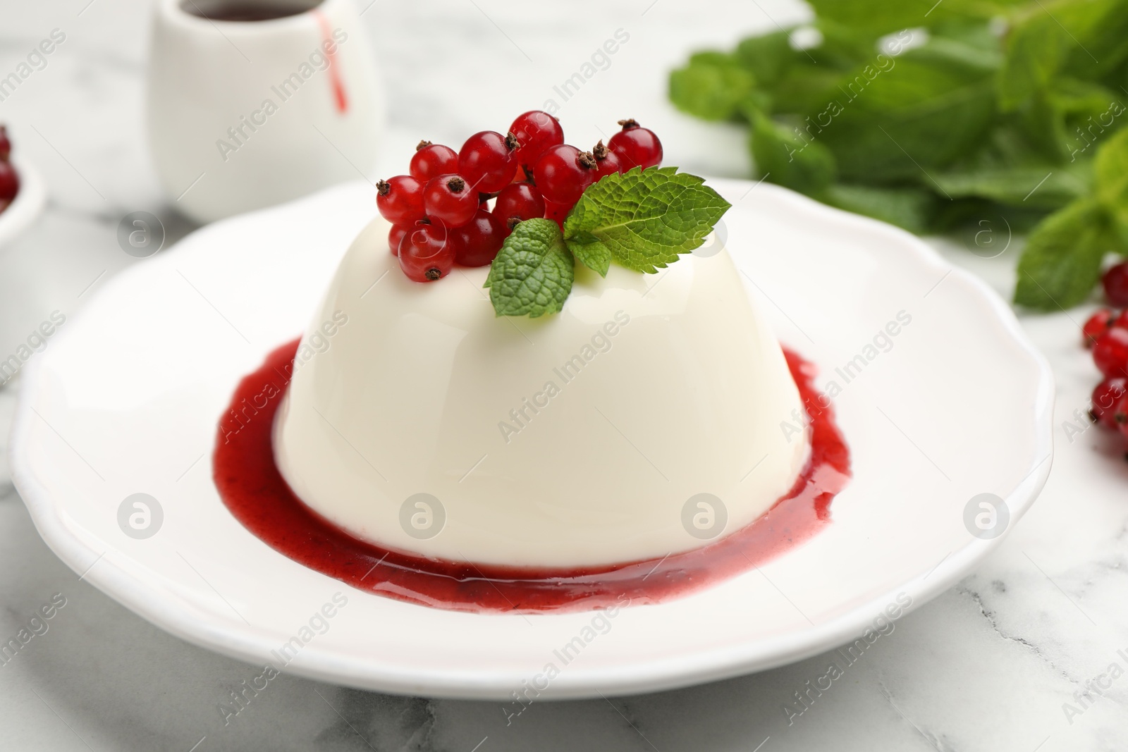 Photo of Delicious panna cotta with red currants, sauce and mint on white marble table, closeup