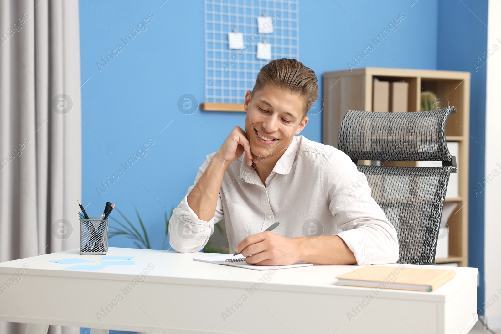 Dream come true. Positive young man thinking about something at desk indoors Photo of Dream come true. Positive young man thinking about something at desk indoors