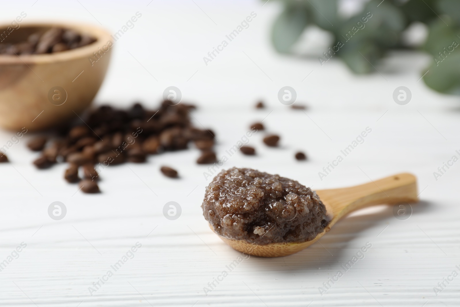 Photo of Natural body scrub in spoon, coffee beans and eucalyptus on white wooden table, closeup. Space for text