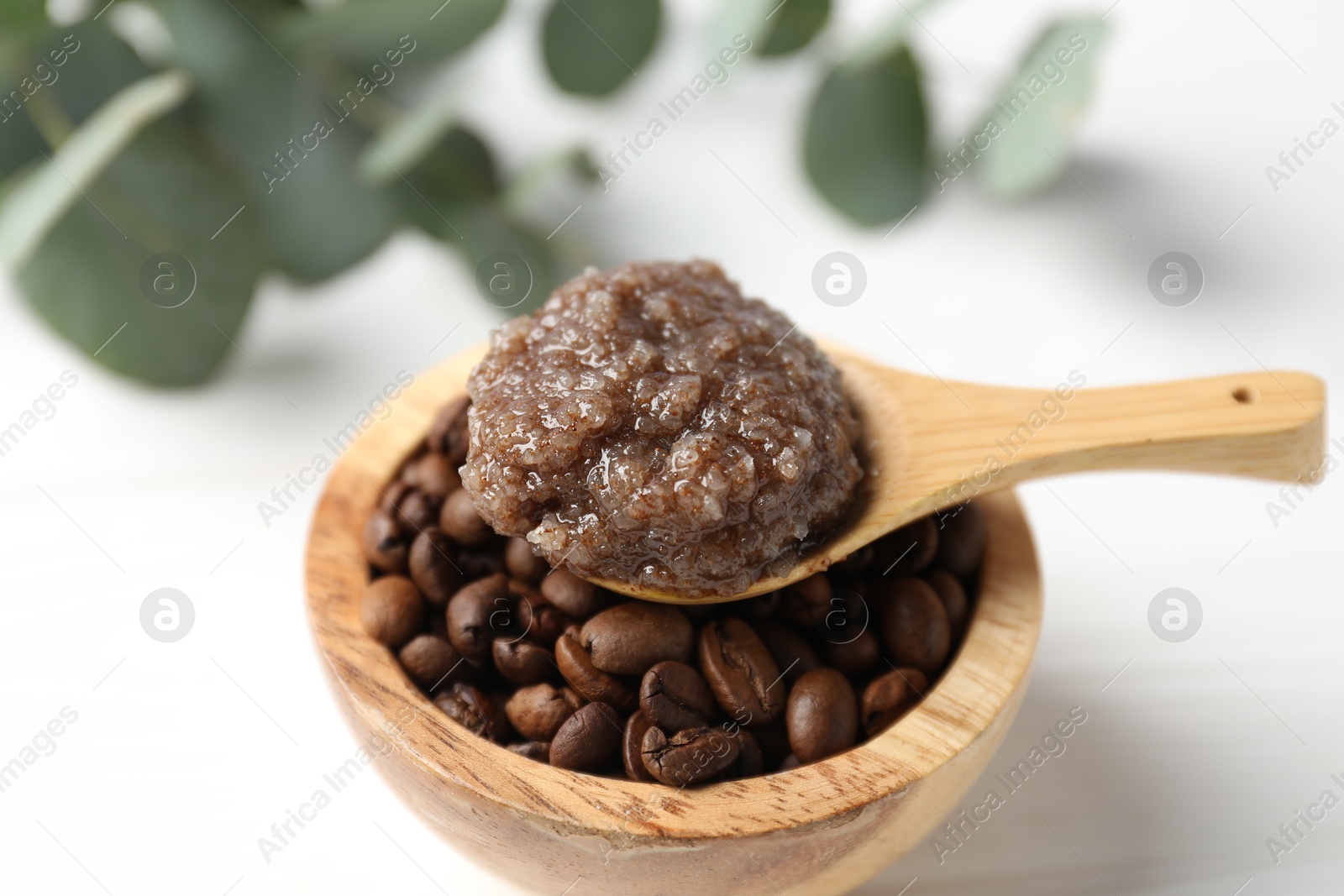 Photo of Natural body scrub in spoon, coffee beans and eucalyptus branches on white table, closeup