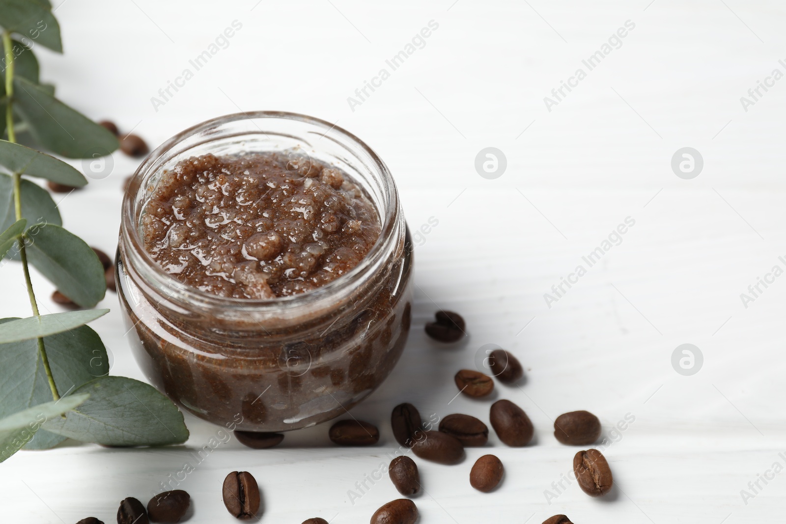 Photo of Natural body scrub in glass jar, coffee beans and eucalyptus branch on white wooden table, closeup. Space for text