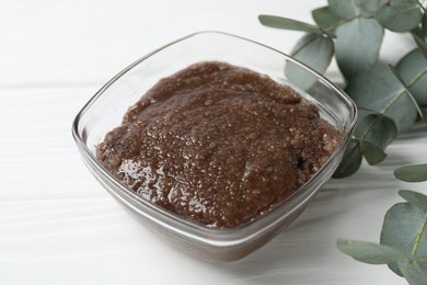 Photo of Natural body scrub in glass bowl and eucalyptus branches on white wooden table, closeup