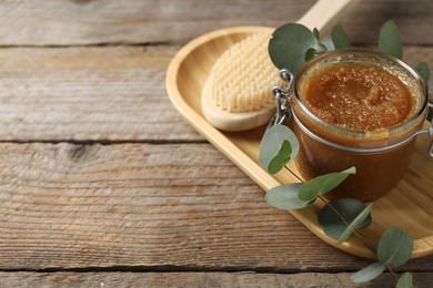 Photo of Natural body scrub in glass jar, brush and eucalyptus branch on wooden table, closeup. Space for text