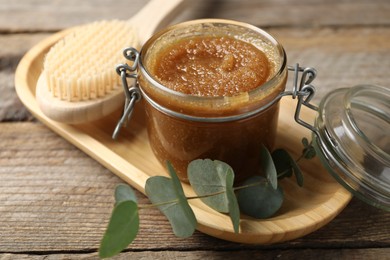 Photo of Natural body scrub in glass jar, brush and eucalyptus branch on wooden table, closeup