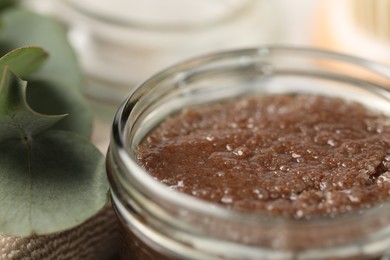 Photo of Natural body scrub in glass jar, eucalyptus leaves and towel on blurred background, closeup