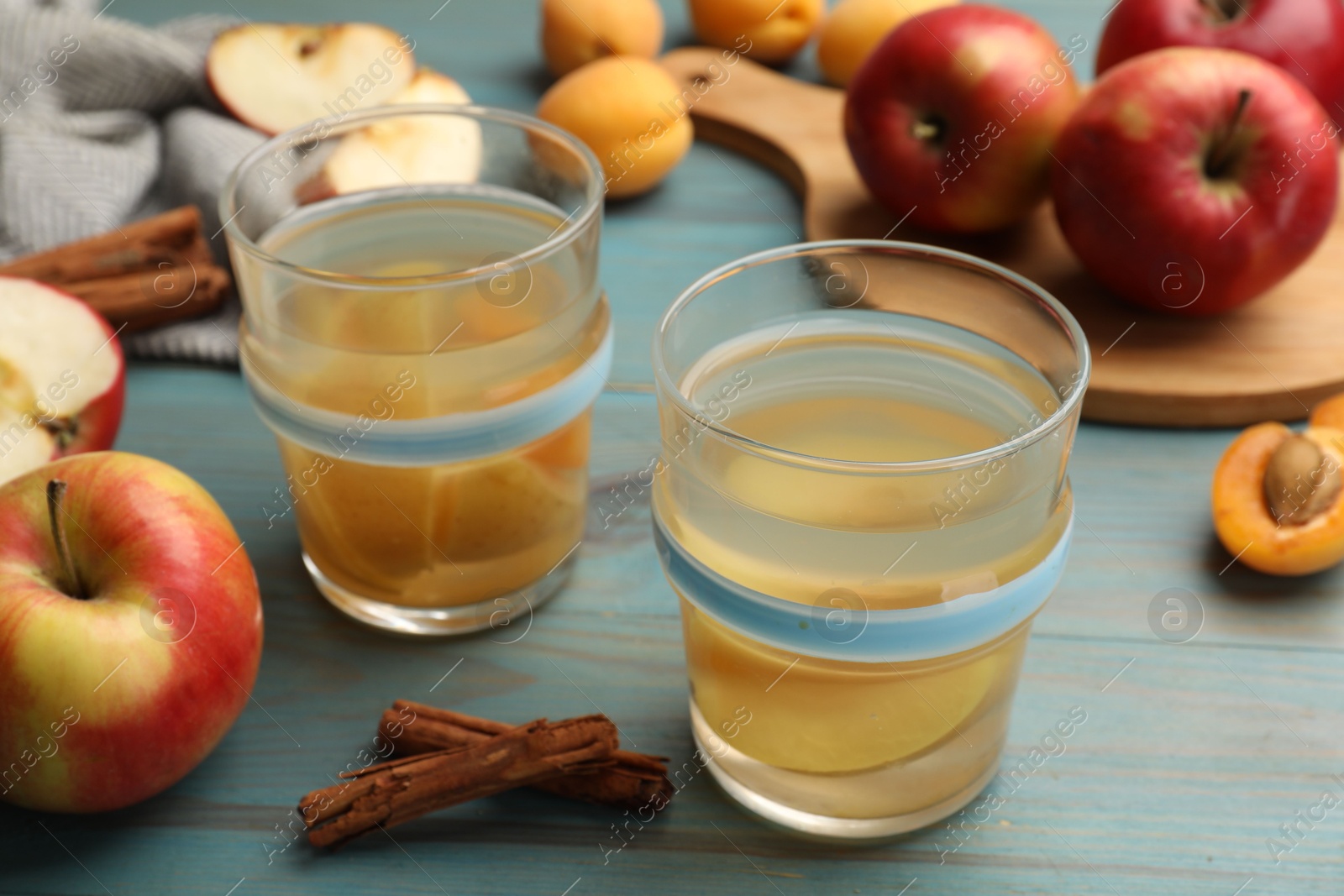 Photo of Tasty compot in glasses, fresh apples, apricots and cinnamon on light blue wooden table, closeup