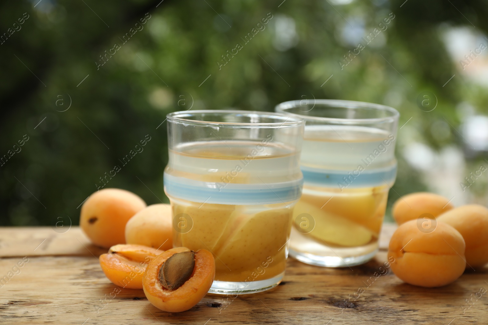 Photo of Tasty compot with apples in glasses and fresh apricots on wooden table outdoors, closeup