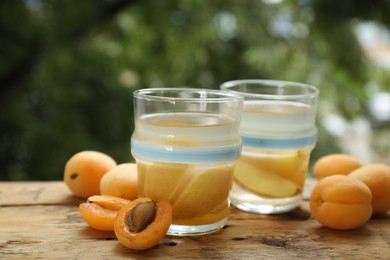 Photo of Tasty compot with apples in glasses and fresh apricots on wooden table outdoors, closeup