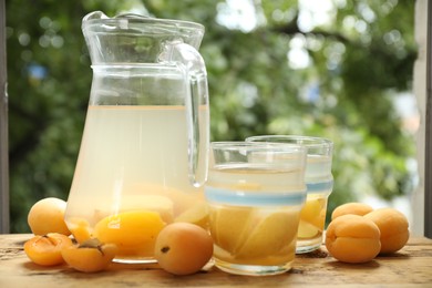 Photo of Tasty compot with apples in glasses, jug and fresh apricots on wooden table outdoors