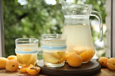 Photo of Tasty compot with apples in glasses, jug and fresh apricots on wooden table outdoors, closeup