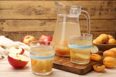 Photo of Tasty compot in glasses, jug, fresh apricots and apples on wooden table, closeup