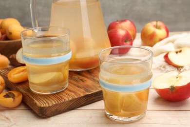 Photo of Tasty compot in glasses, jug, fresh apricots and apples on wooden table, closeup