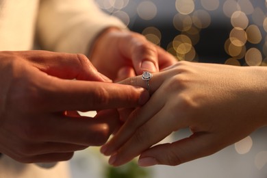 Photo of Making proposal. Man putting engagement ring on his girlfriend's finger indoors, closeup. Bokeh effect