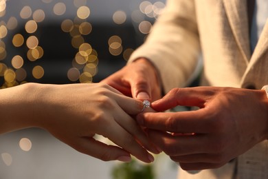 Photo of Making proposal. Man putting engagement ring on his girlfriend's finger indoors, closeup. Bokeh effect