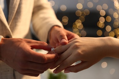 Photo of Making proposal. Man putting engagement ring on his girlfriend's finger indoors, closeup. Bokeh effect