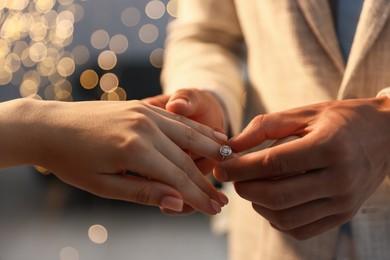 Photo of Making proposal. Man putting engagement ring on his girlfriend's finger indoors, closeup. Bokeh effect
