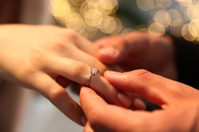 Photo of Making proposal. Man putting engagement ring on his girlfriend's finger indoors, closeup. Bokeh effect