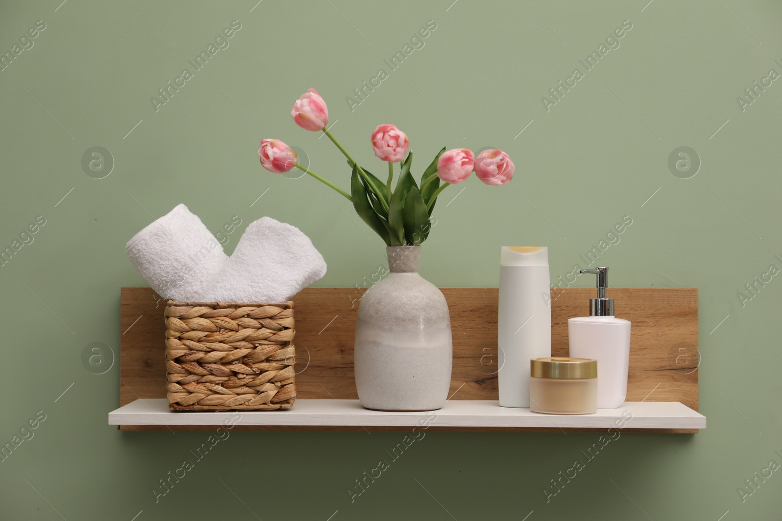Photo of White wooden shelf with vase of pink tulips, towels and cosmetic products on light green wall