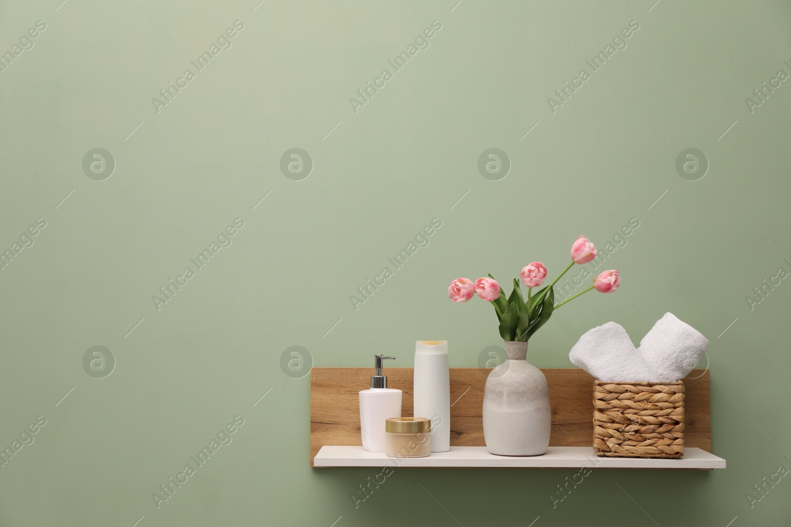 Photo of White wooden shelf with vase of pink tulips, towels and cosmetic products on light green wall, space for text