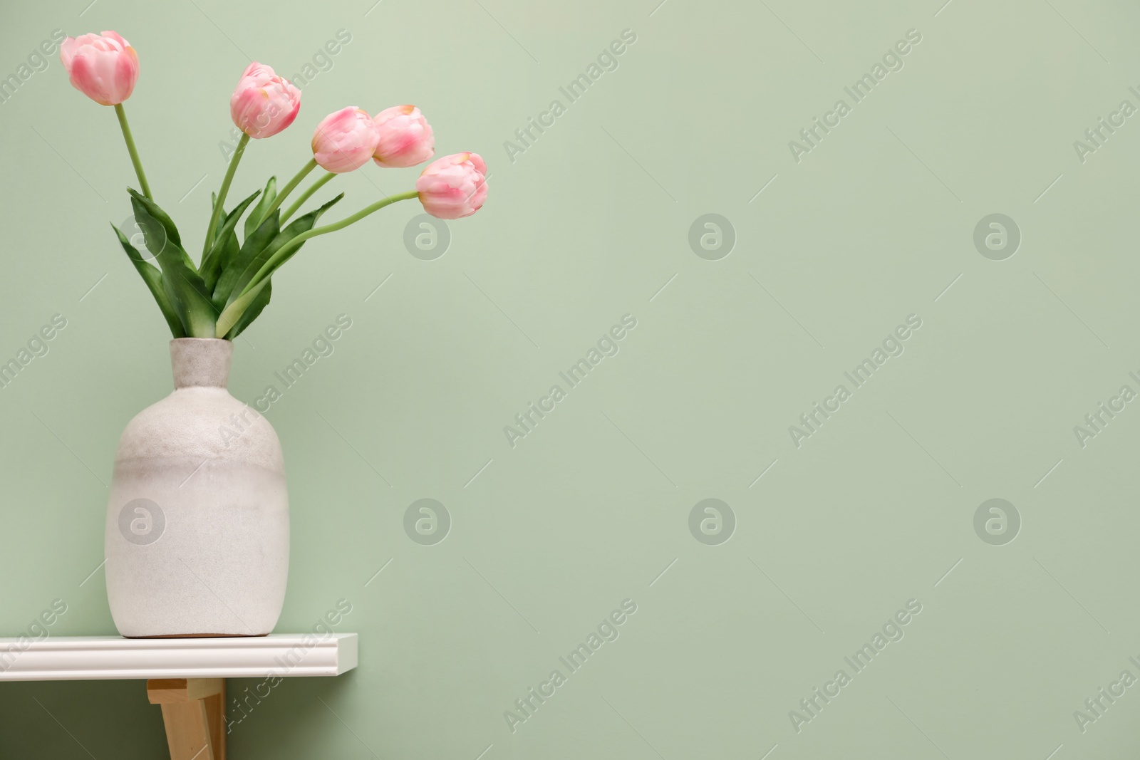 Photo of White shelf with vase of pink tulips on light green wall, space for text