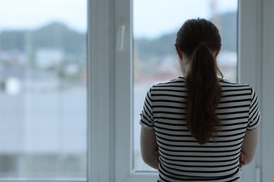 Photo of Sad young woman near window at home, back view. Space for text