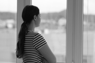 Photo of Sad young woman near window at home, back view and space for text. Black and white effect