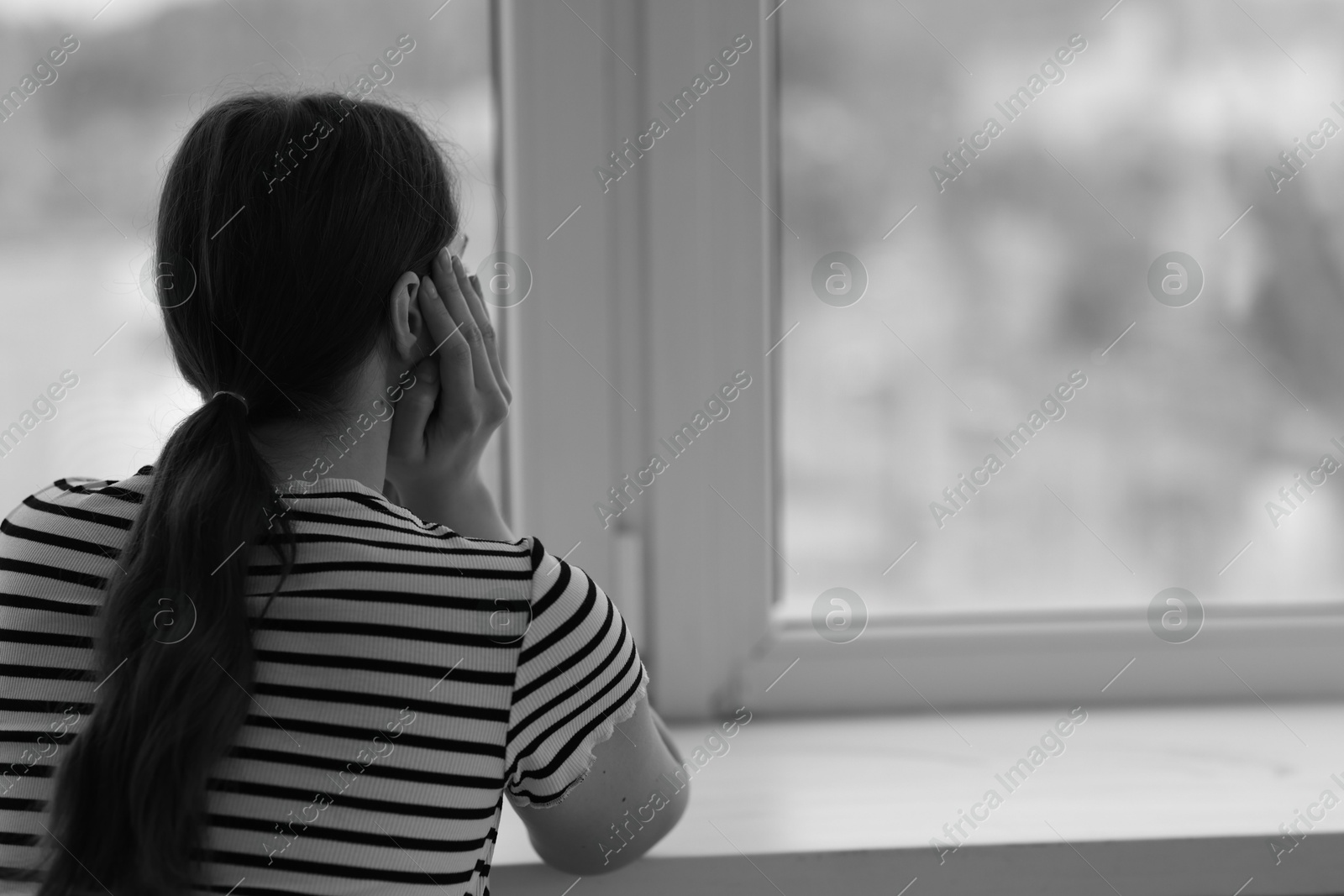 Photo of Sad young woman near window at home, back view and space for text. Black and white effect