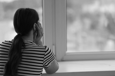 Photo of Sad young woman near window at home, back view and space for text. Black and white effect