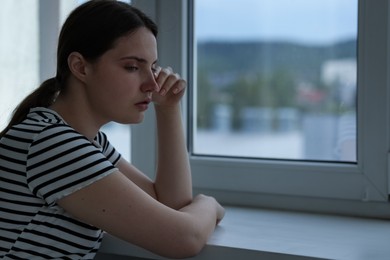Photo of Sad young woman near window at home, space for text