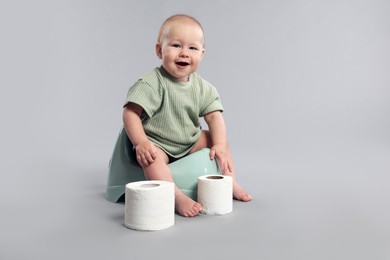 Photo of Cute little baby sitting on potty and rolls of toilet paper against light grey background