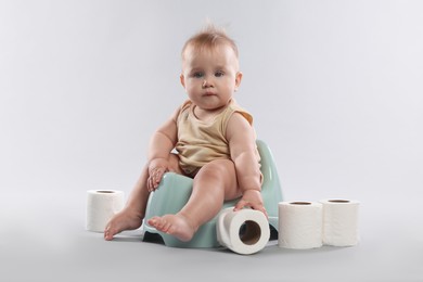 Photo of Cute little baby sitting on potty and rolls of toilet paper against light grey background