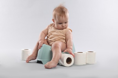 Photo of Cute little baby sitting on potty and rolls of toilet paper against light grey background